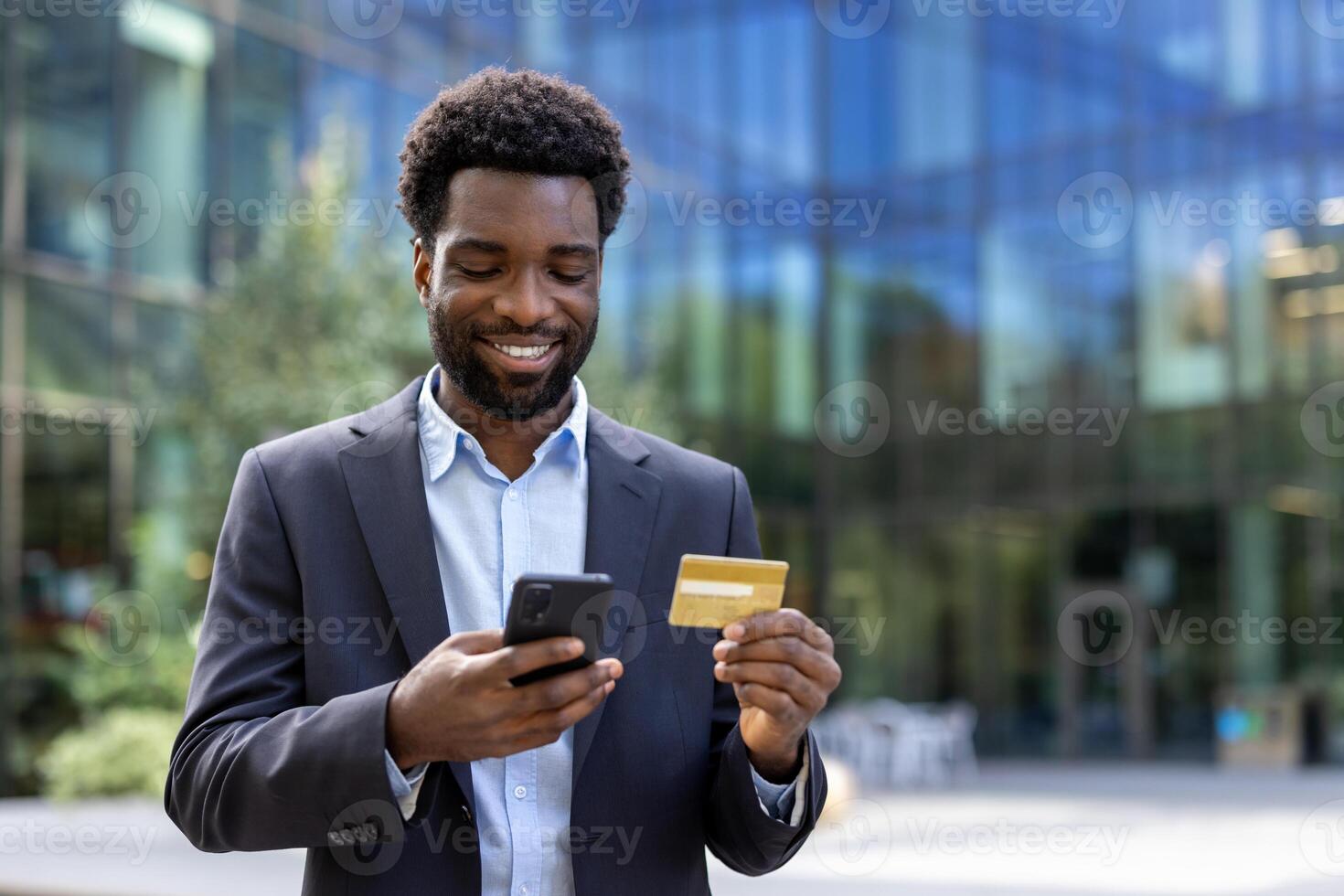 Successful businessman walking outside office building with phone in hand and bank credit card. Man in business suit shopping online in online store and booking services remotely. photo