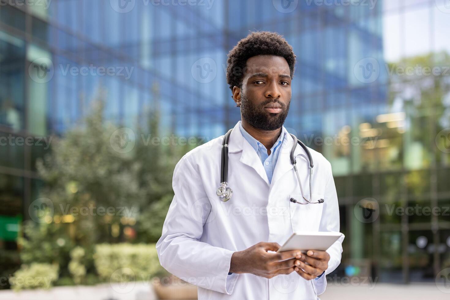 Portrait of a young confident and serious doctor. A man with a tablet computer in his hands and in a white medical coat looks focused at the camera while walking outside the clinic office. photo