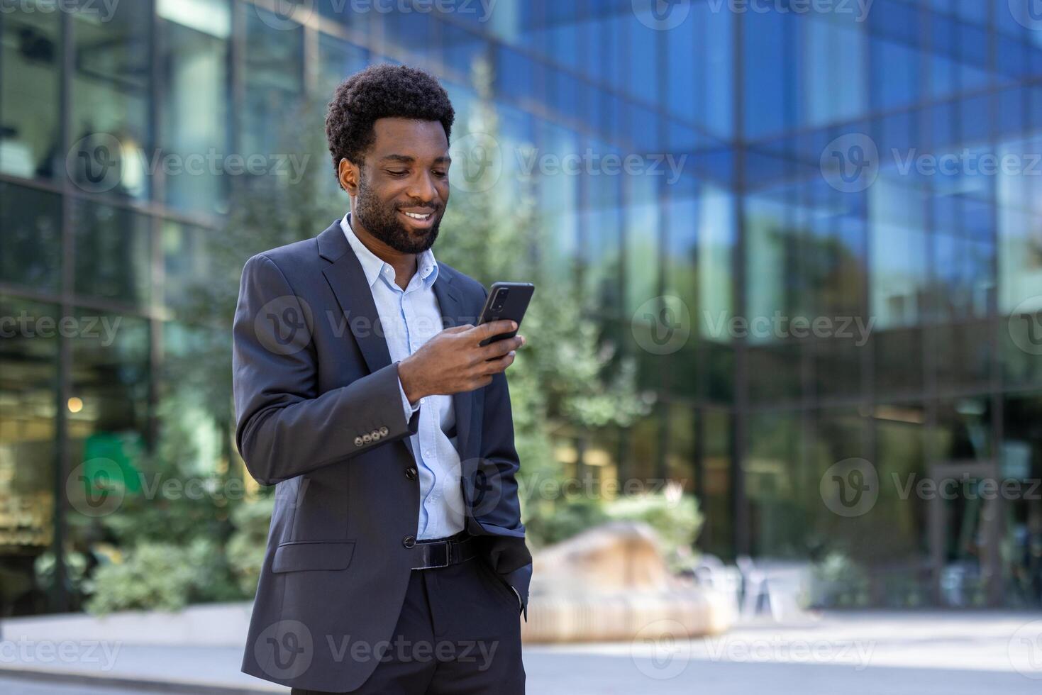 Successful businessman walking through the city with phone in hand. Joyful African American man in business suit using smartphone app, browsing social media, and typing text message. photo