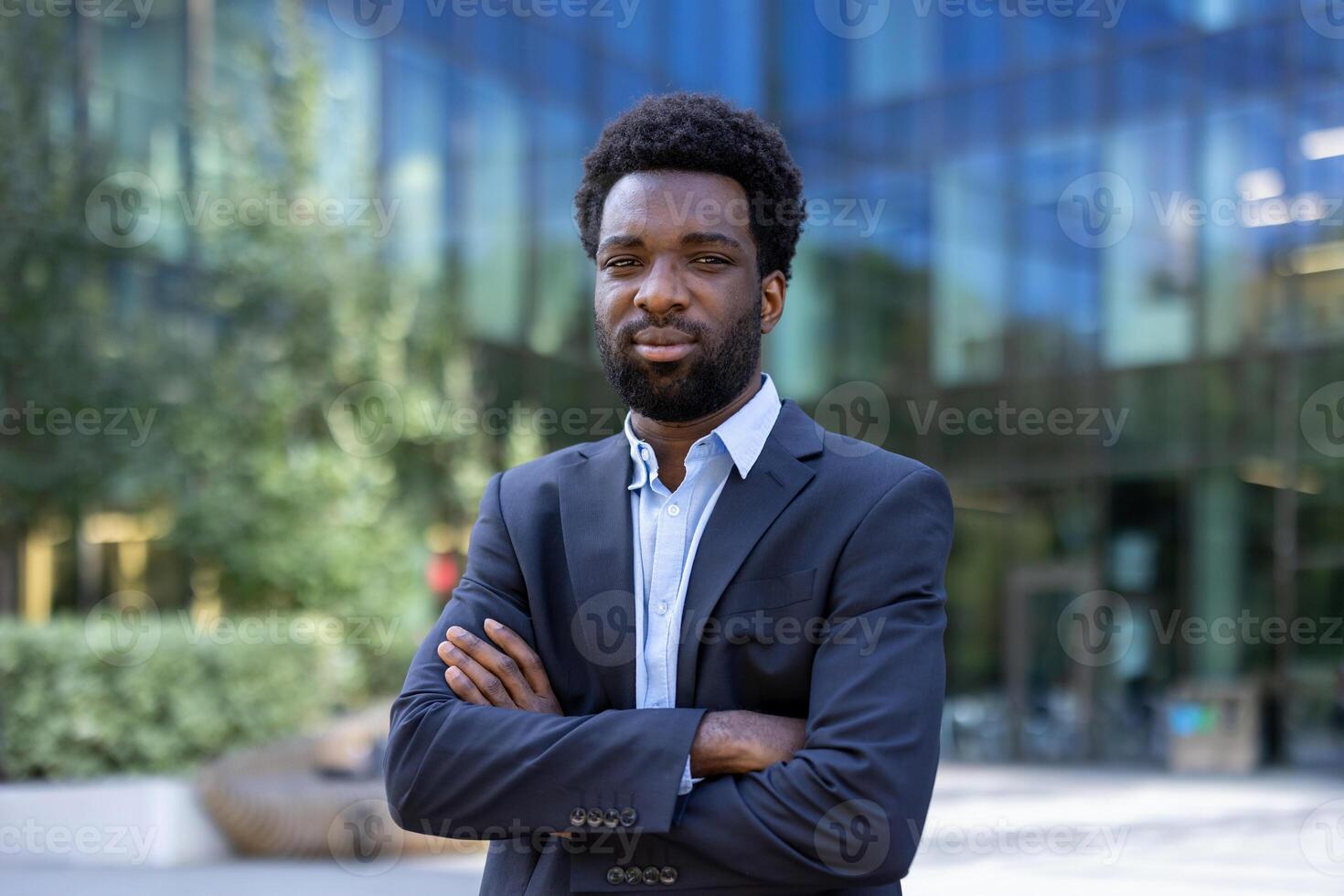 A confident Black businessman stands with crossed arms in front of a modern office building with blurred greenery. photo
