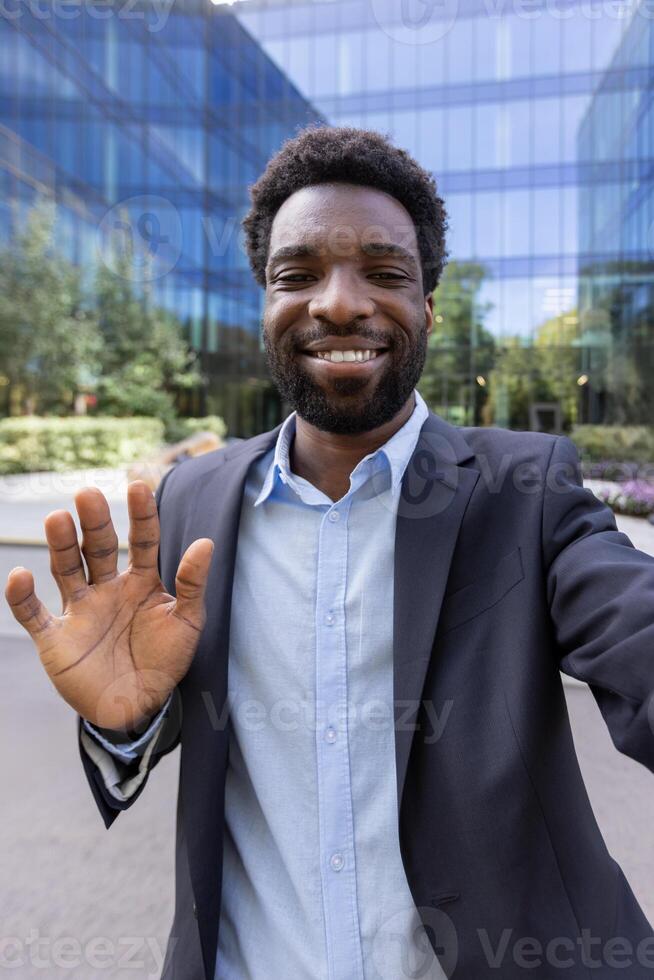 A young, handsome man in a blazer smiles and waves to the camera during a mobile call in front of a modern office building photo