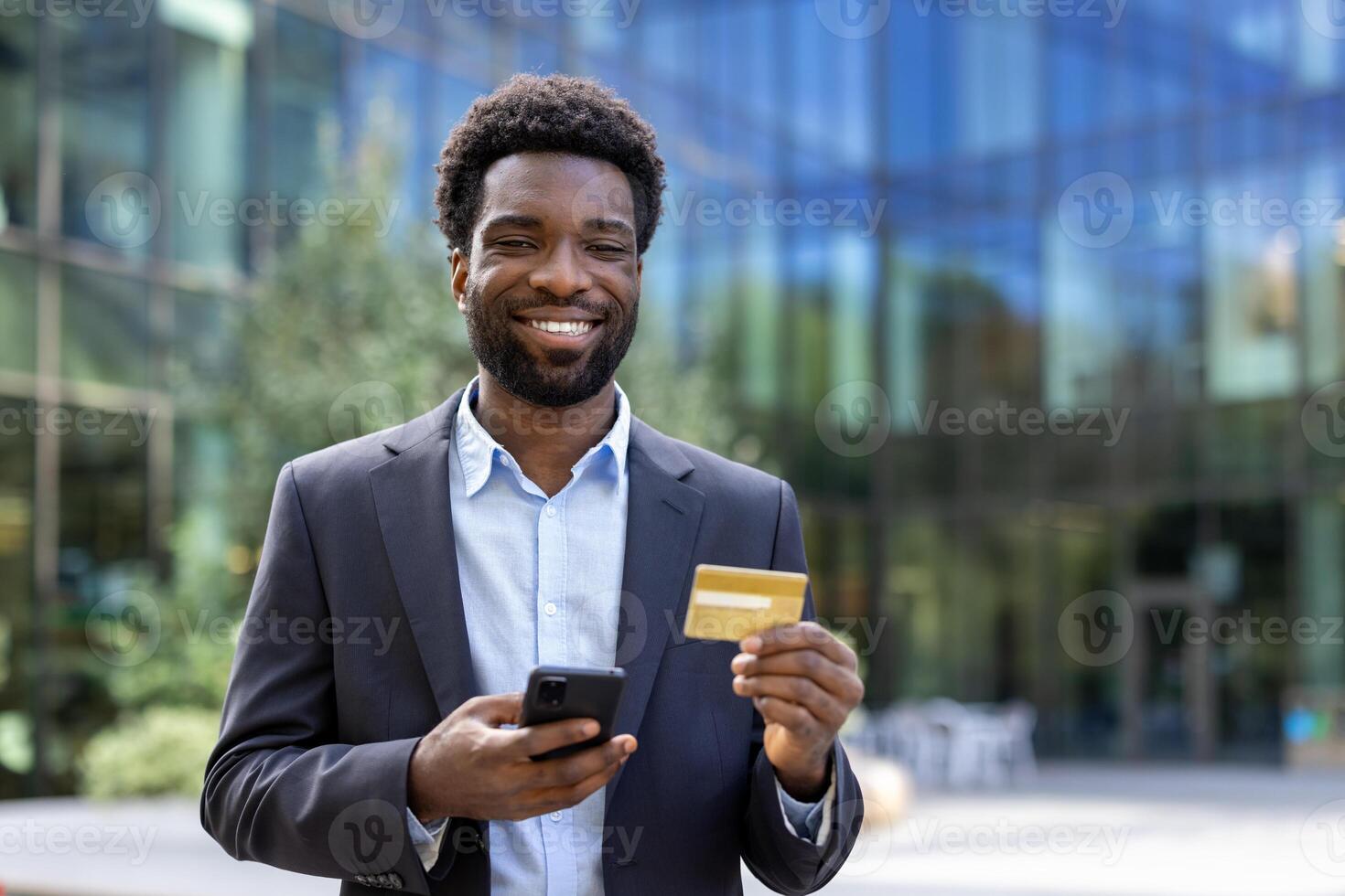 Joyful businessman with bank credit card and phone outside office building. Man smiling and looking at camera, satisfied with online shopping and money transfer remotely. photo