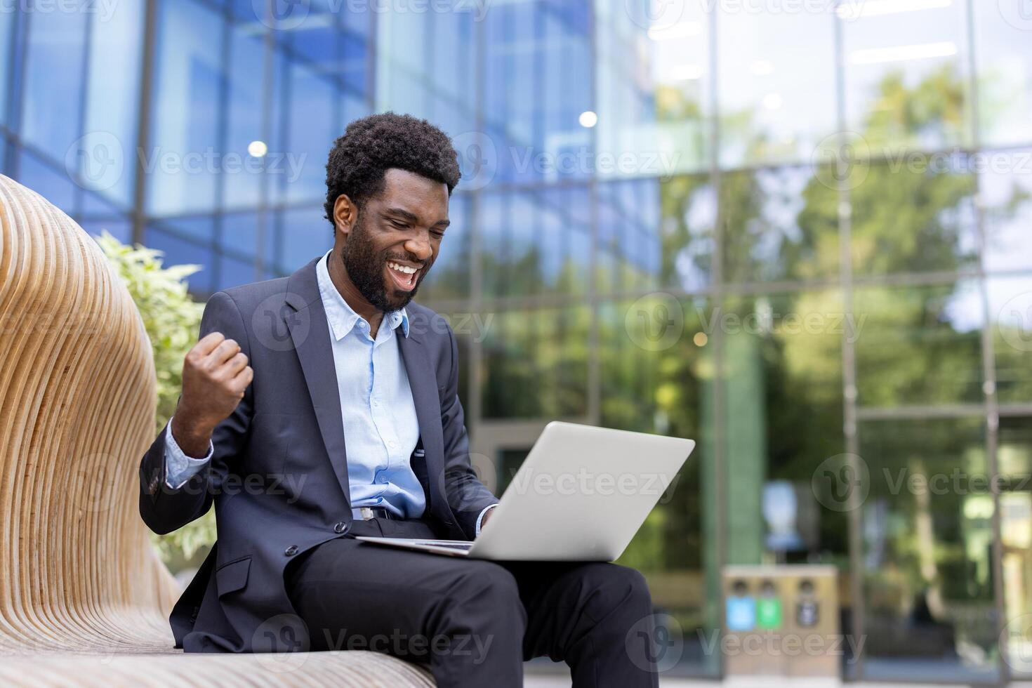 A cheerful businessman celebrates success while working remotely on his laptop outside an office building on a bench, expressing joy and excitement. photo