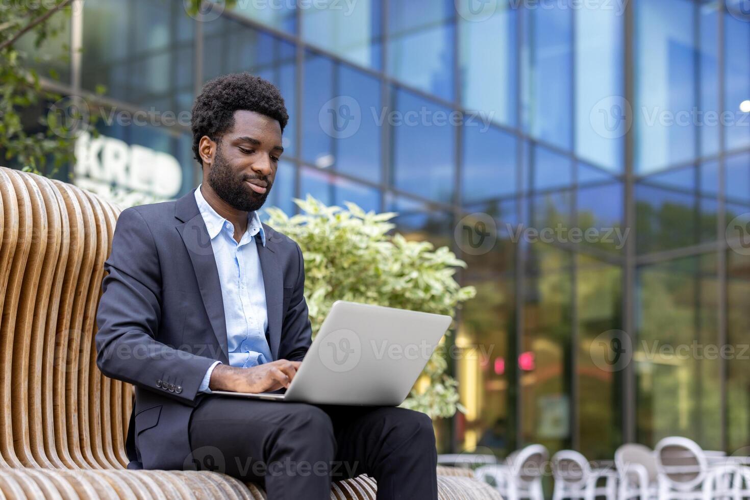 A young professional works remotely, utilizing his laptop while seated on a modern bench with an office building as a backdrop. photo