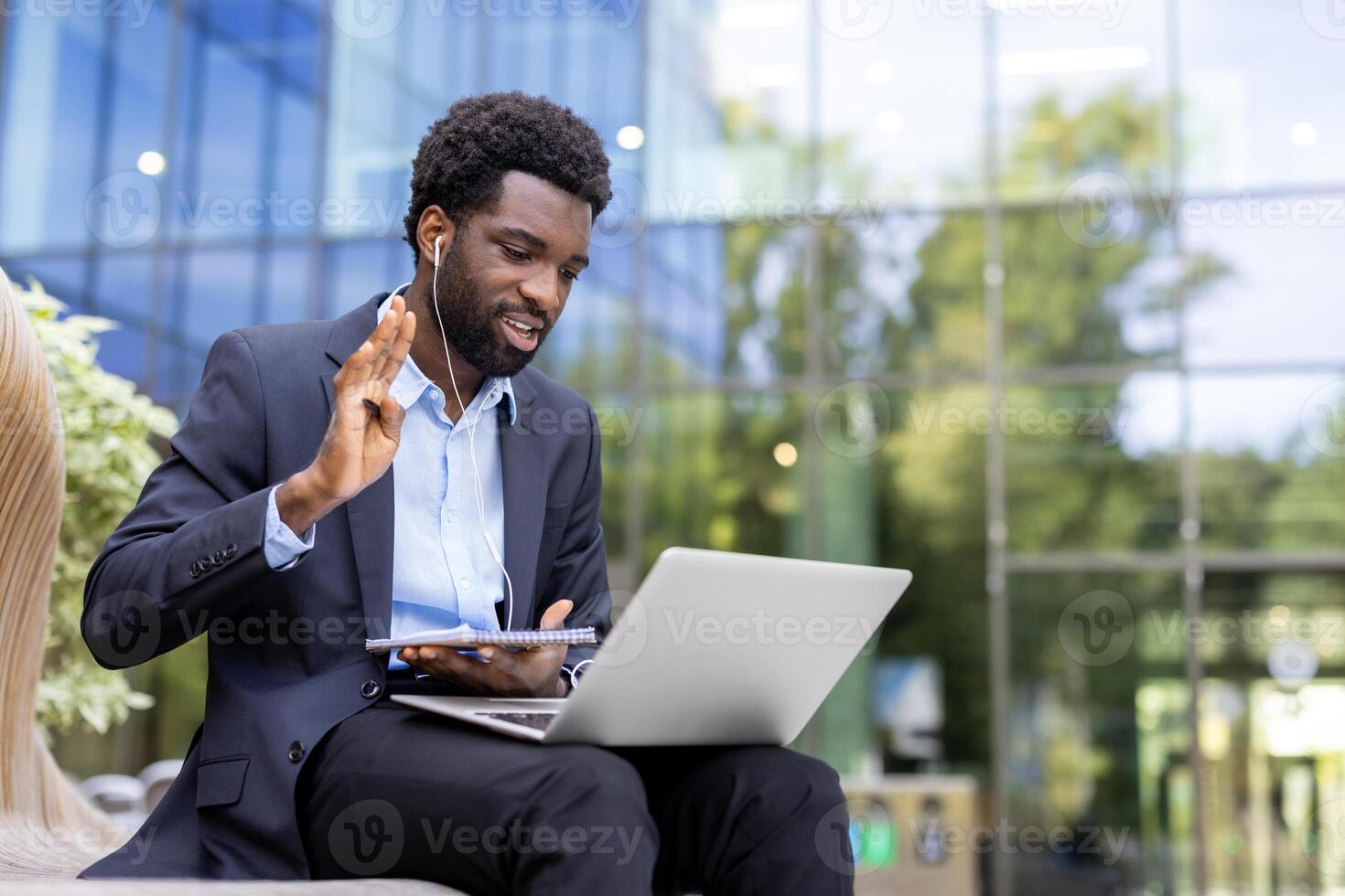 A confident African-American businessman attends a virtual meeting on his laptop outdoors, greeting colleagues and making notes in a notebook. photo