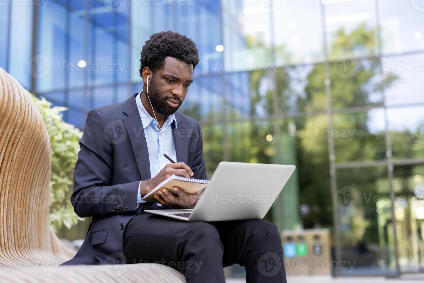 Focused businessman in a suit works outdoors on a laptop while jotting notes, wearing earbuds against a modern building backdrop. photo