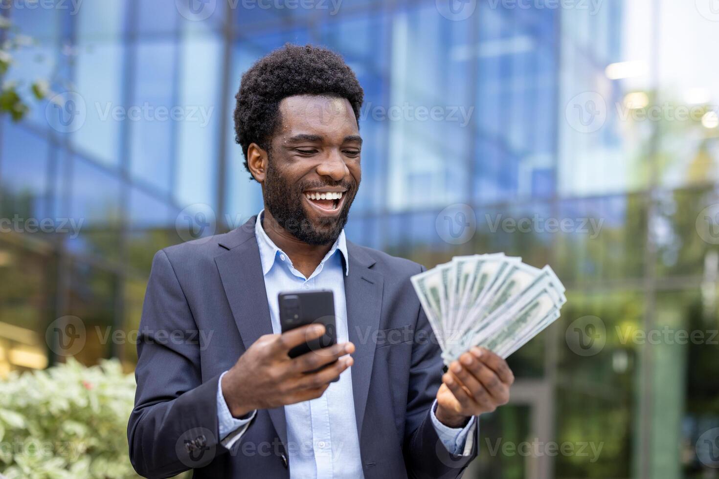 A young successful businessman celebrates holding a fan of cash while looking at his smartphone, outdoors in front of an office building. photo