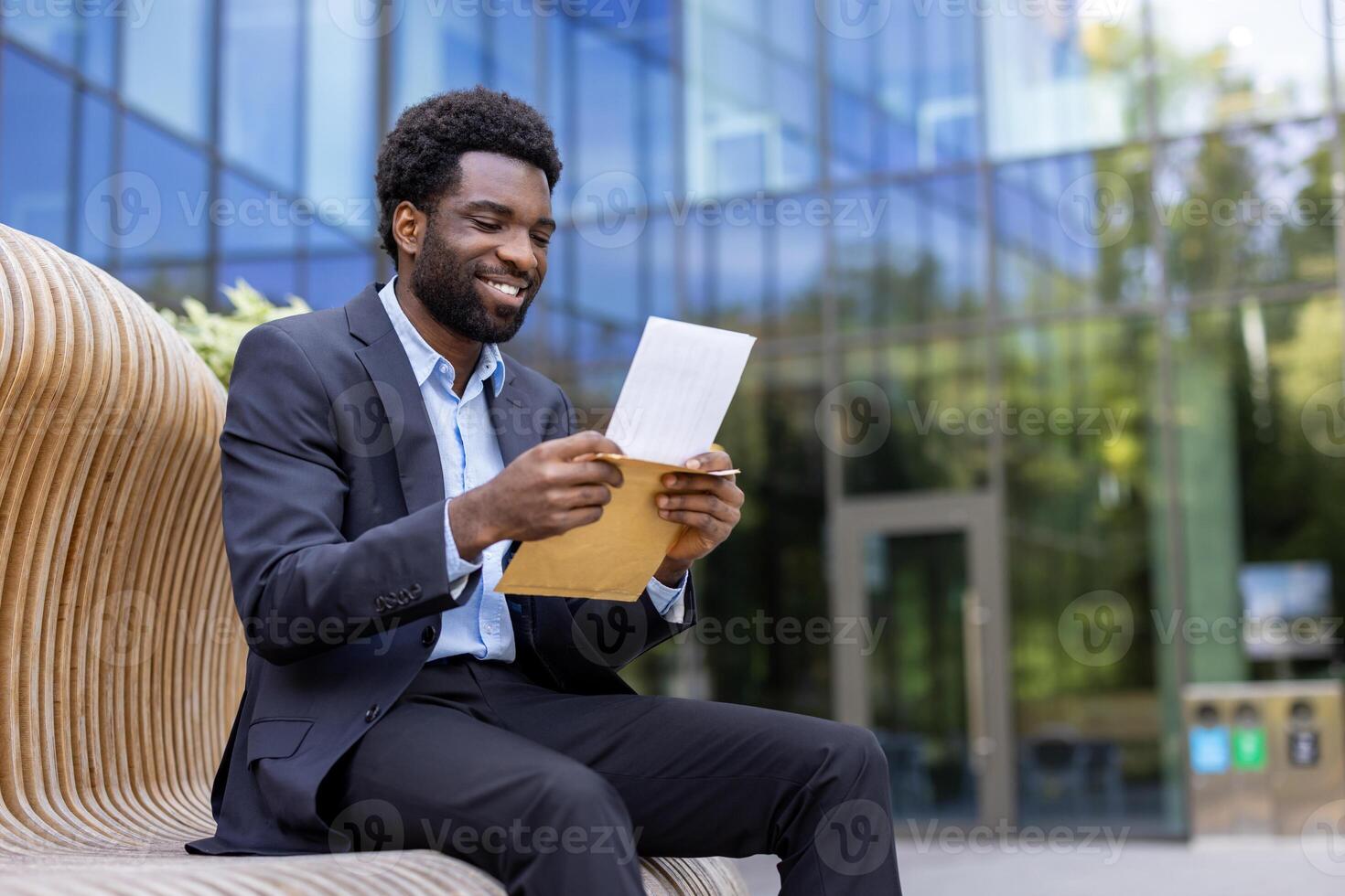 An african american businessman in a suit sits outside, smiling while reading a document in an envelope, happy. The setting is a modern office environment. photo