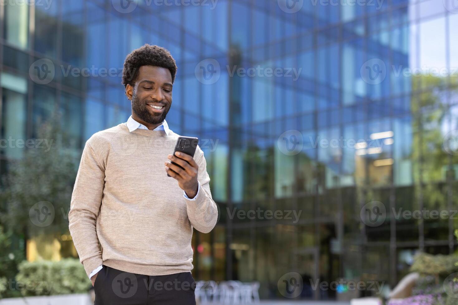 An attractive Black man smiles while looking at his smartphone, standing outdoors in front of a modern office building. He appears happy. photo