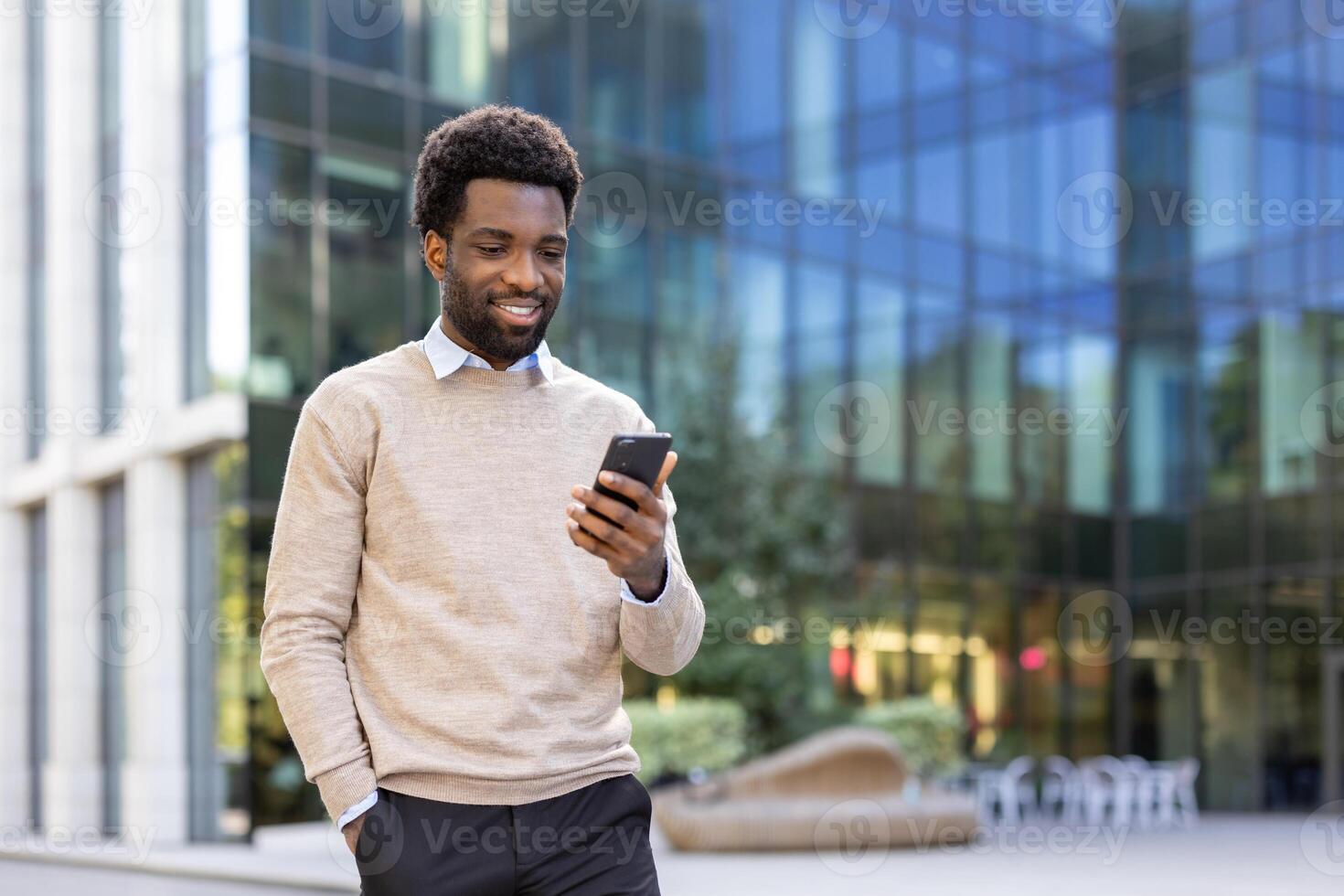 An african-american man stands in front of a glass building, looking at his phone with a pleasant expression. It's a sunny day. photo