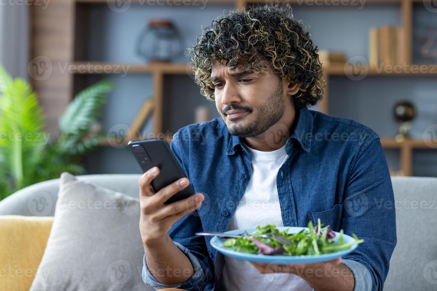 A young man with curly hair checks his smartphone while holding a plate of salad at home on the couch during lunchtime. photo
