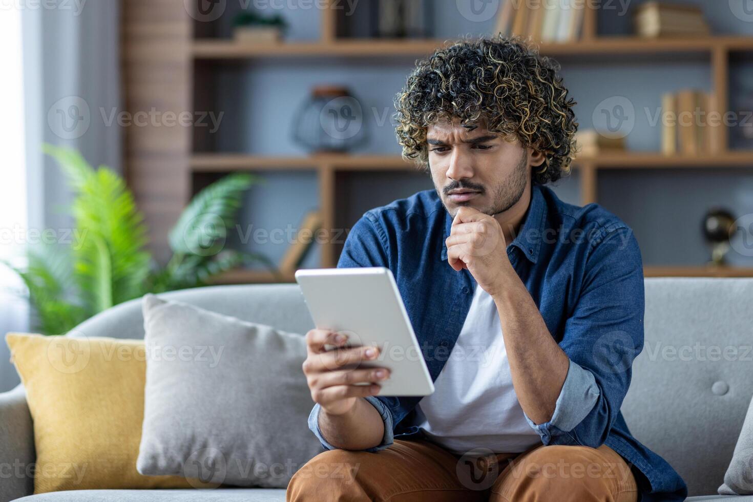 Serious thinking man using tablet computer reading e-book while sitting on sofa in living room at home. photo