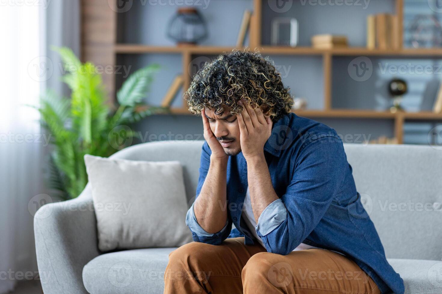 Stressed man with curly hair sitting on a couch, experiencing a bad headache in a modern interior. He is overwhelmed with negative emotions. photo