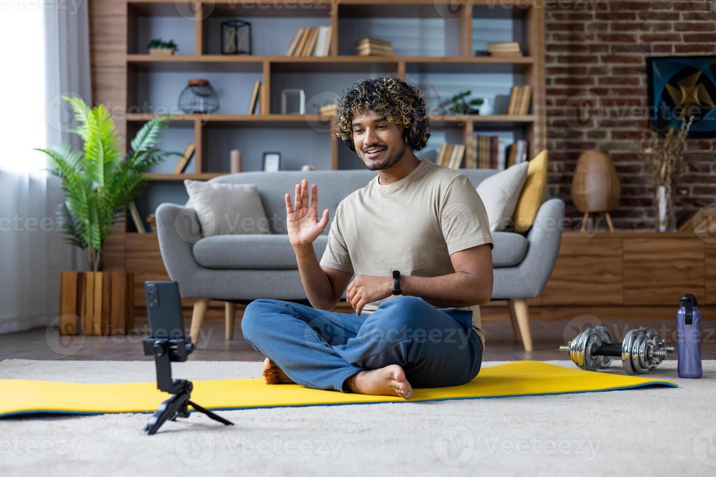 A smiling man waves while conferencing at home during his fitness routine. The scene includes a yoga mat and dumbbells. photo