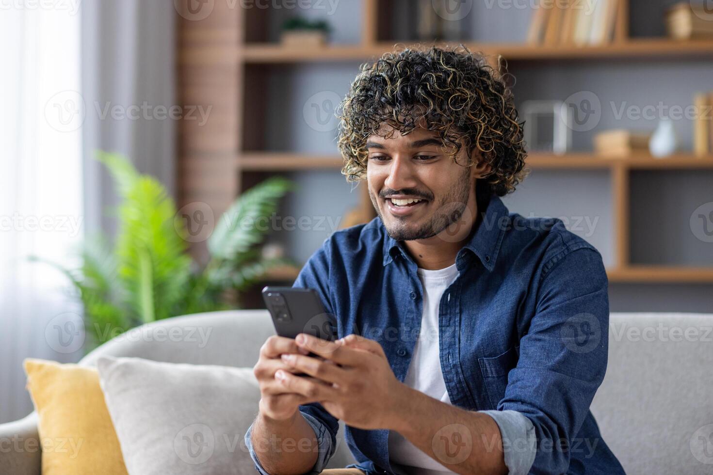 A young man with curly hair sits on a sofa, using a smartphone and smiling widely. photo