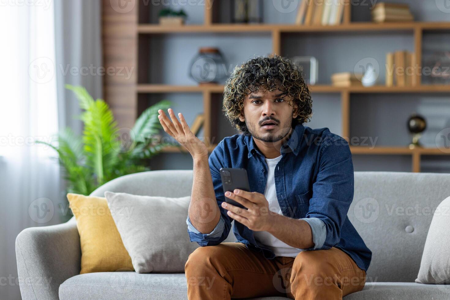 A young man looks confused while holding his smartphone, questioning a message or notification, seated on a couch in a home setting. photo