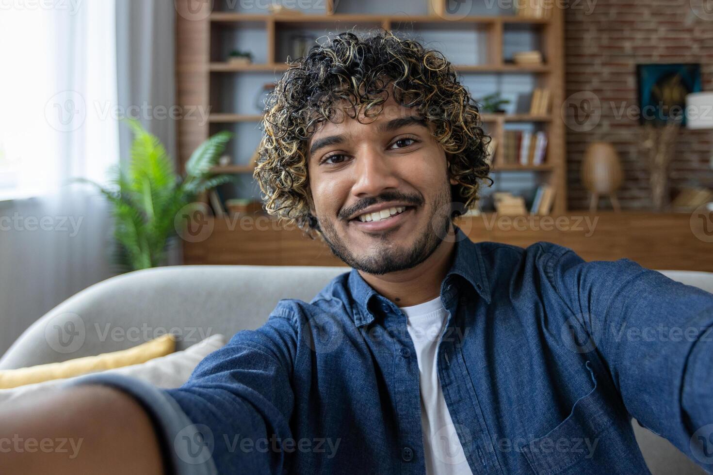 A smiling man with curly hair takes a selfie at home, casually dressed in a blue shirt, with a glimpse of his cozy living space in the background. photo