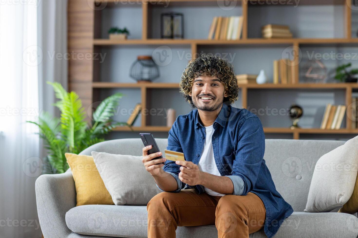 Young man with curly hair is sitting on the sofa at home shopping online, using a phone and a credit card for the payment transaction. photo