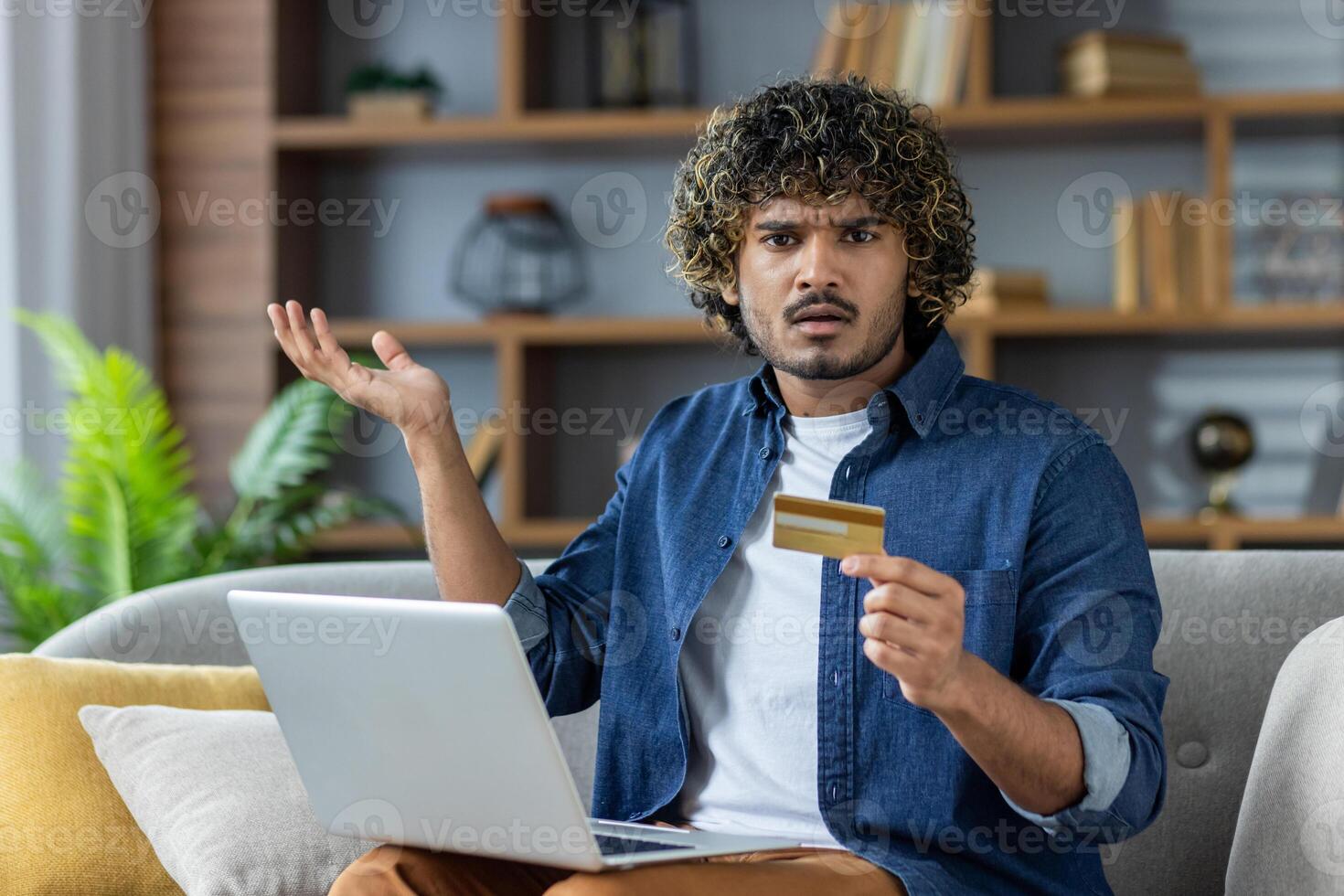 Young man looks confused while holding a credit card and using a laptop at home, likely encountering online payment issues. photo