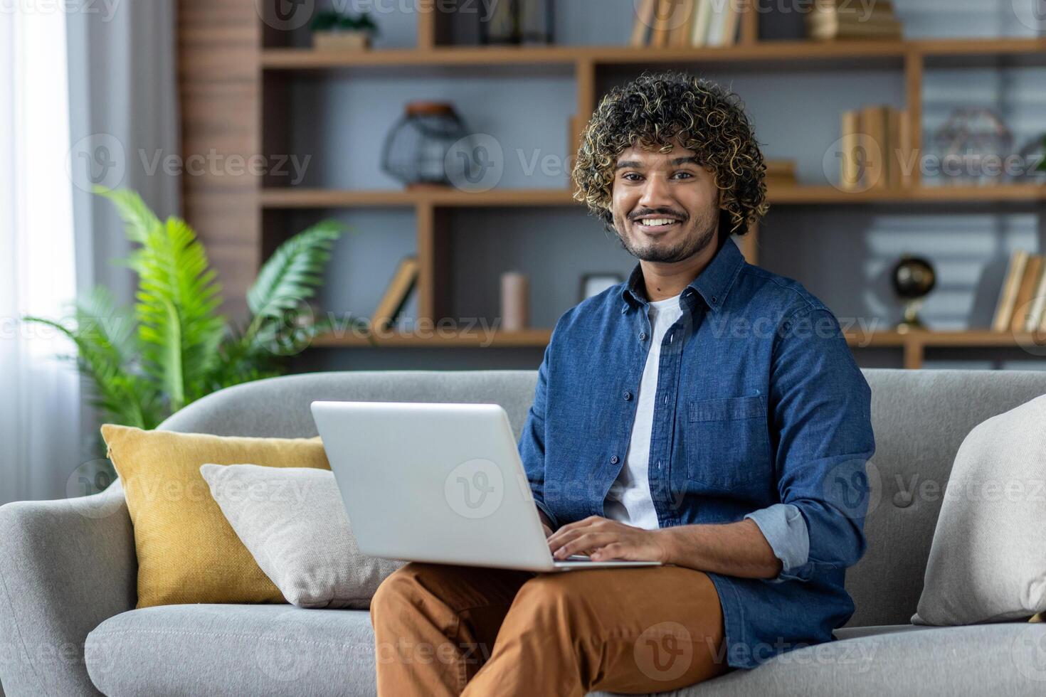 A young man with curly hair smiles while working on a laptop while seated on a gray sofa at home. The background features a cozy living room. photo