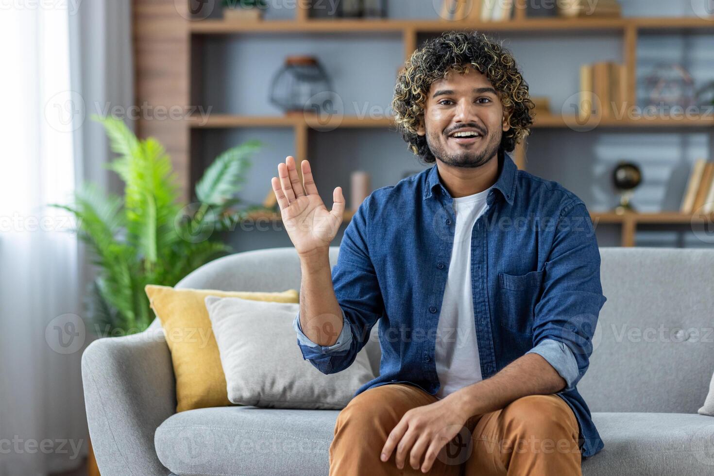 A cheerful young man with curly hair is waving with a smile while sitting on a sofa in his living room, looking into the camera. photo