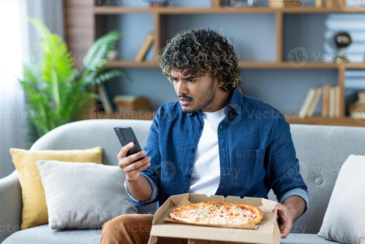 A man looks at his phone with a confused expression while holding a pizza box, possibly dealing with an order issue photo