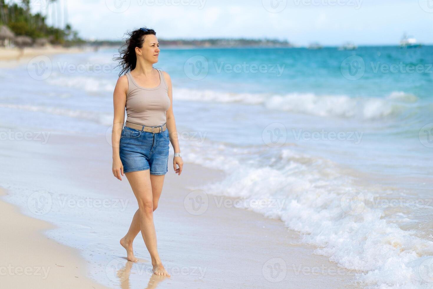 Woman walking barefoot along a serene beach during a sunny day with gentle waves in the background photo
