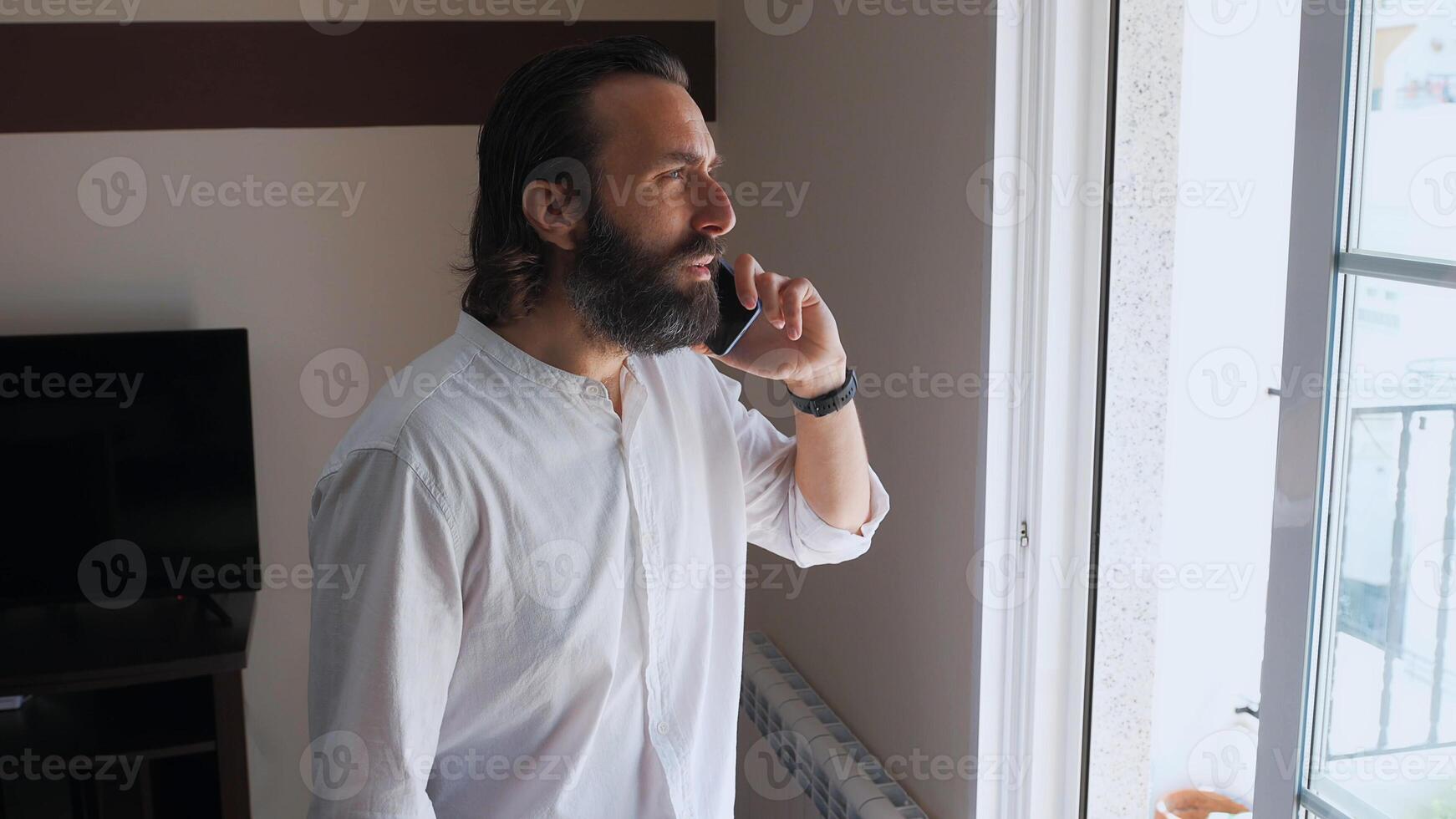 Confident Bearded Man Talking on Smartphone Near a Window photo