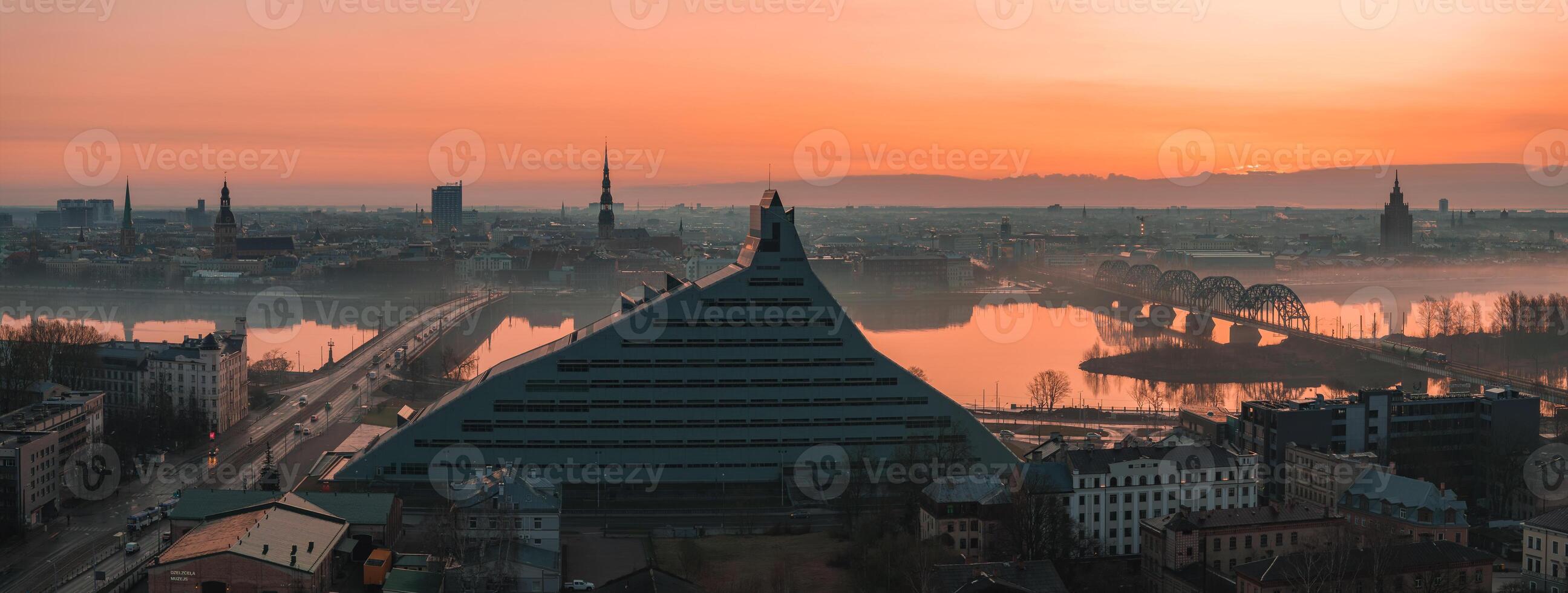 Morning view of Riga, Latvia, featuring the National Library, historic church spires, the Daugava River, and the arched railway bridge in soft mist. photo