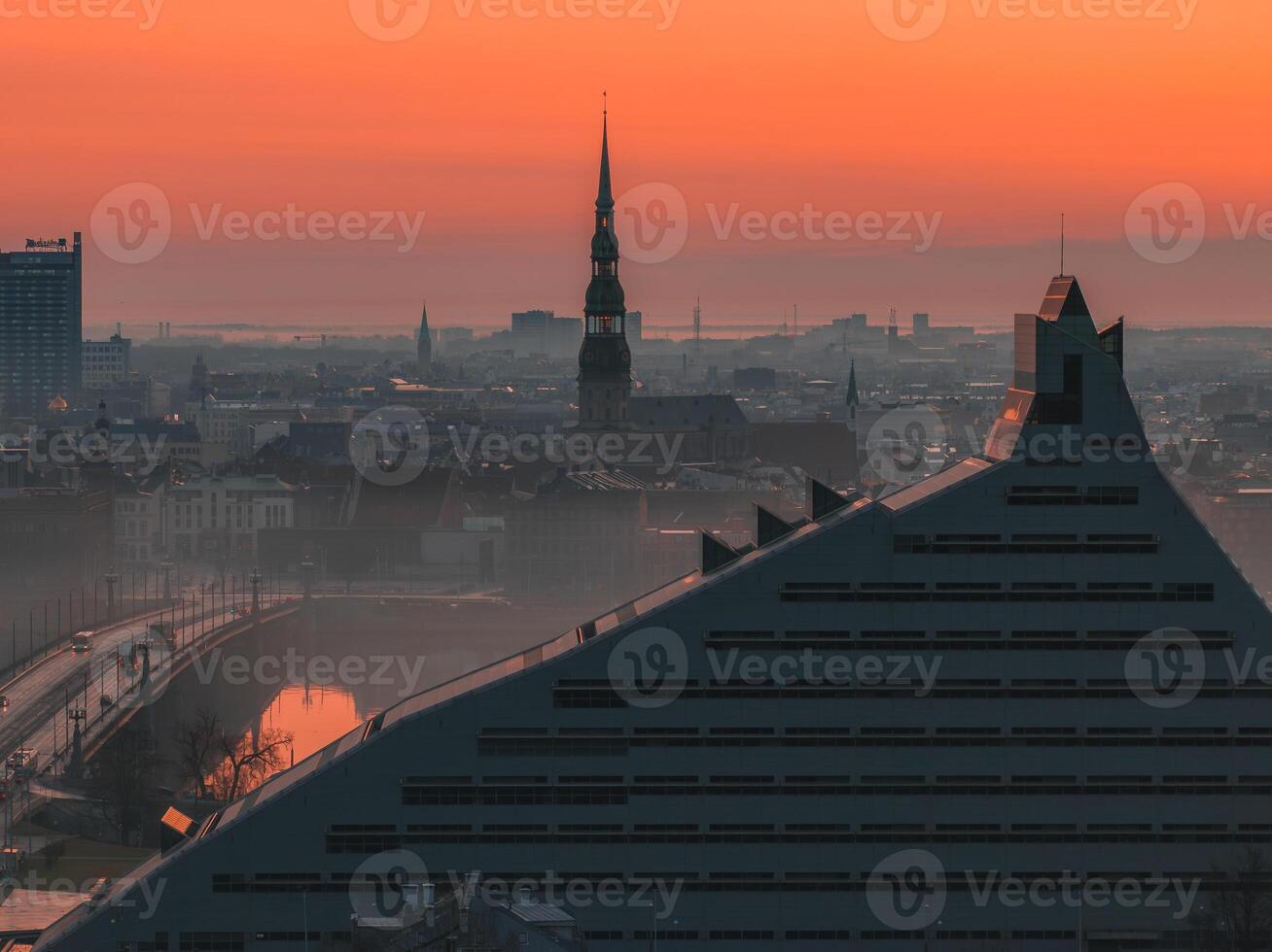 Serene sunrise over Riga, Latvia, featuring St. Peter's Church spire, historic cityscape, modern Latvian National Library, bridge, and waterway. photo