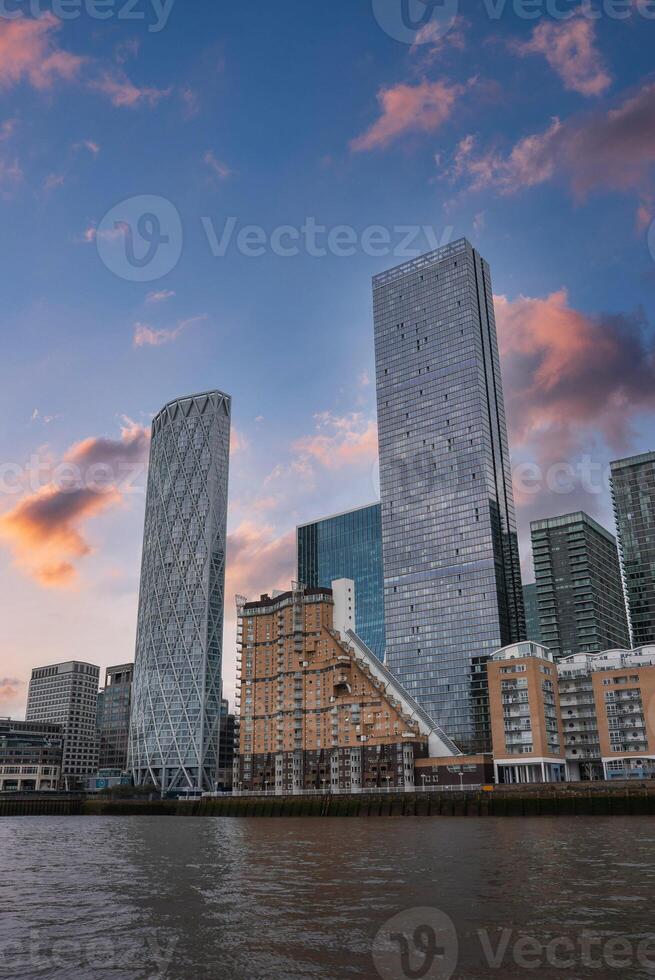 View of London's skyline at sunset with the Thames River, Newfoundland building, and One Canada Square in the Canary Wharf area under a pink orange sky. photo
