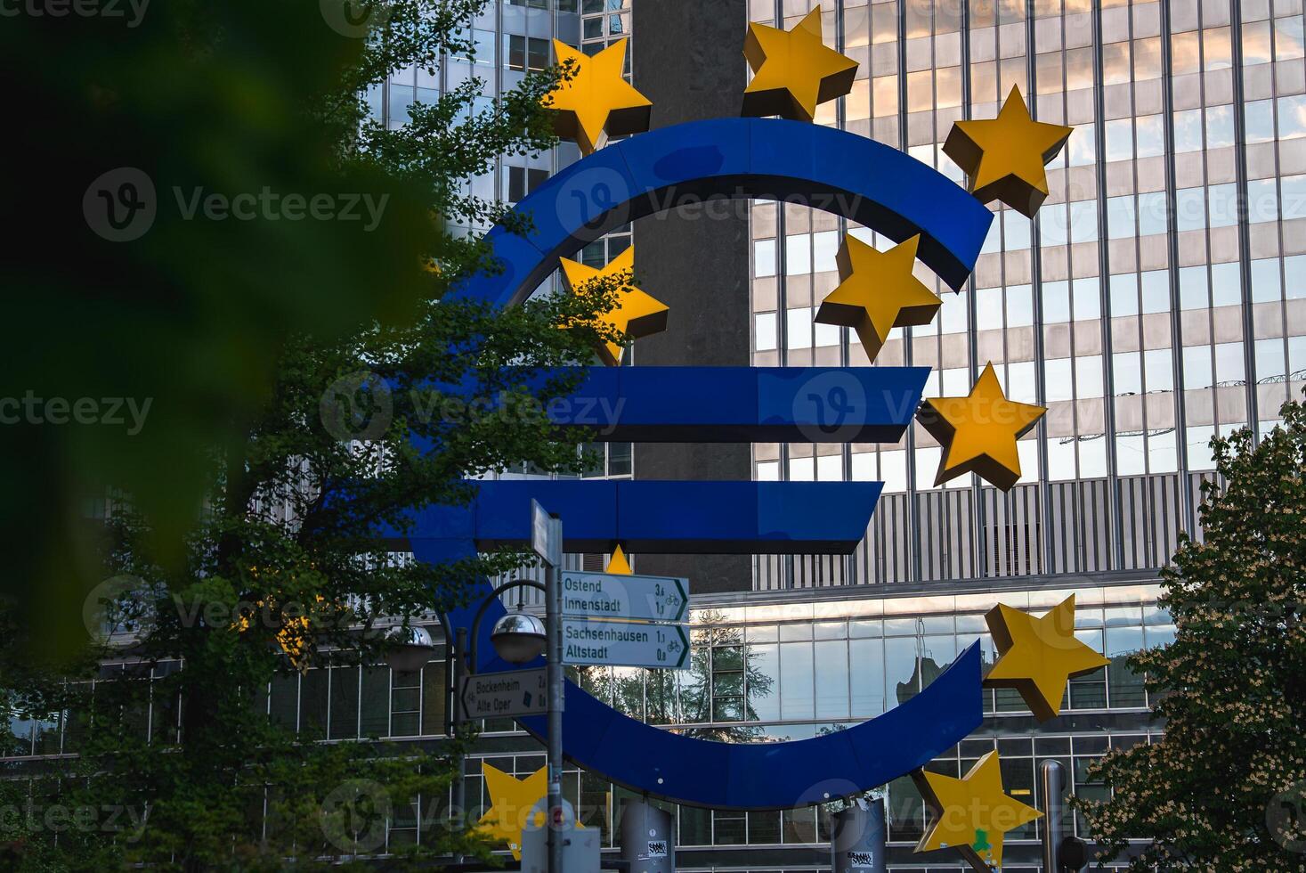 The iconic Euro sculpture with yellow stars stands near the European Central Bank in Frankfurt, Germany, surrounded by modern skyscrapers under a cloudy sky. photo
