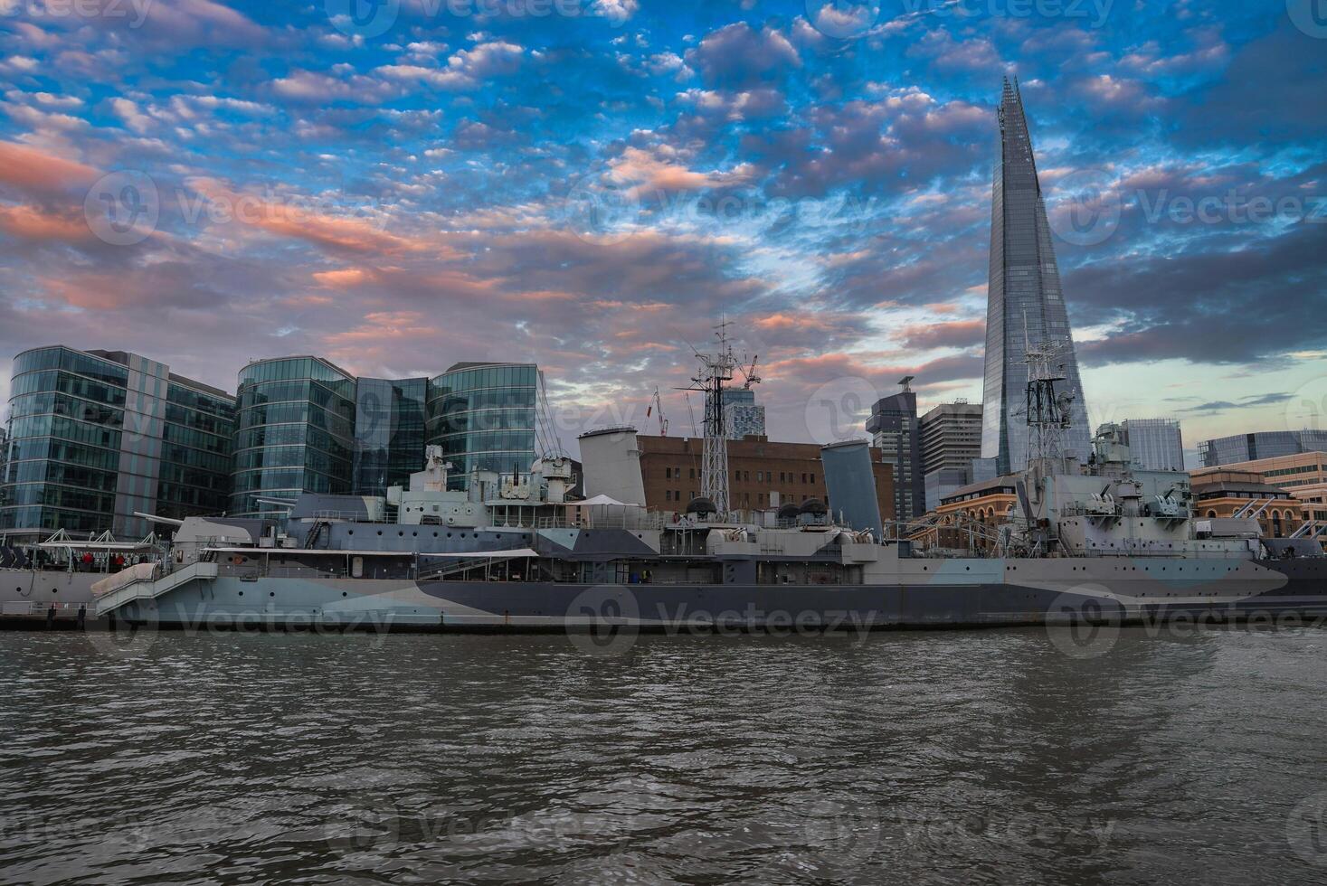 View of London's skyline with the Shard skyscraper, HMS Belfast on the River Thames, and modern glass buildings under a cloud streaked sky. photo