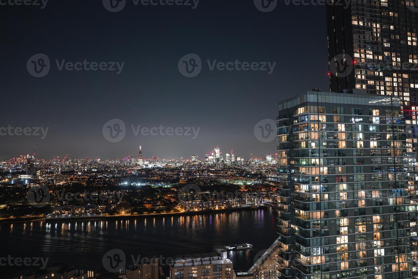 Illuminated London skyline at night featuring The Shard, modern high rises, and the River Thames reflecting city lights in the foreground. photo