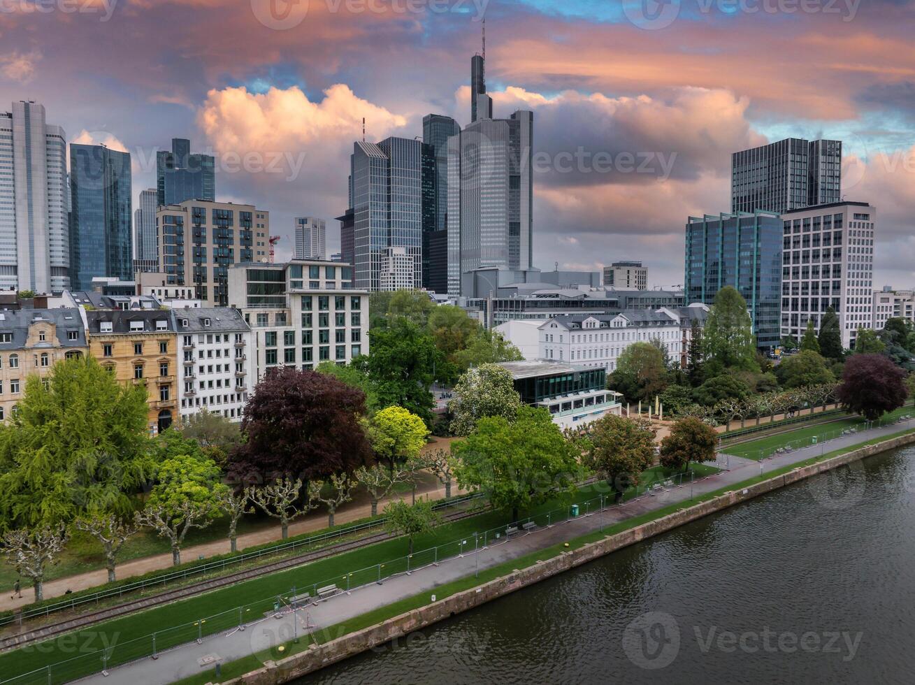 Aerial view of Frankfurt, Germany, featuring the Commerzbank Tower, riverside park greenery, and vibrant orange and pink sunset skies over the Main River. photo