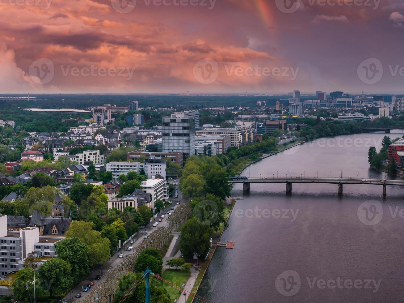 Aerial image of Frankfurt, Germany, featuring the Main River, a bridge, urban skyline, lush greenery, and a vibrant sunset with a faint rainbow. photo
