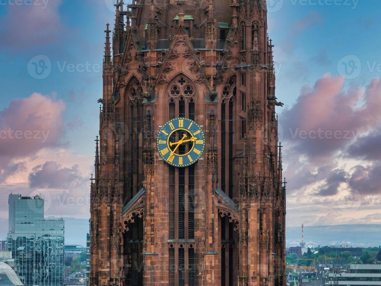 The Gothic style Frankfurt Cathedral tower stands out with intricate details and a gold accented clock, set against modern buildings and a pink blue sky. photo