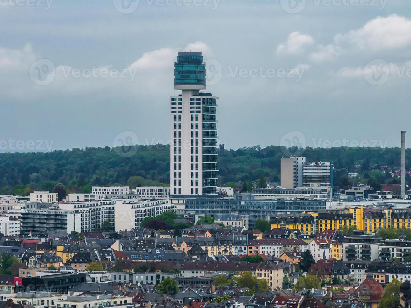 Aerial view of Frankfurt, Germany, highlighting a modern high rise with a cylindrical glass top, surrounded by urban housing, greenery, and a cloudy sky. photo