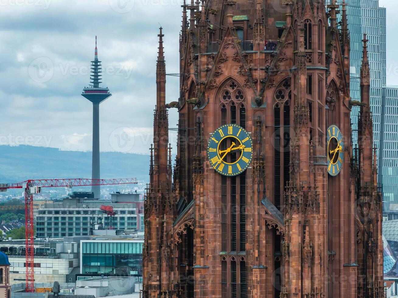 The Frankfurt Cathedral's Gothic spires and clock face stand out, with the Europaturm in the background. Cranes and modern buildings complete the urban scene. photo
