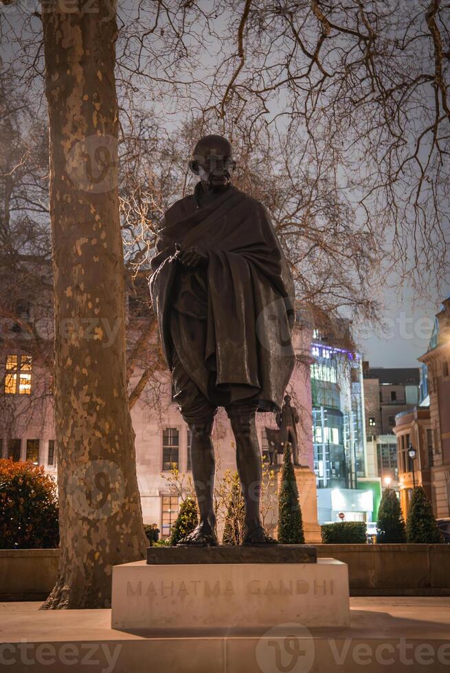 A statue of Mahatma Gandhi on a pedestal surrounded by bare trees, illuminated buildings, and streetlights in a London outdoor setting at night. photo