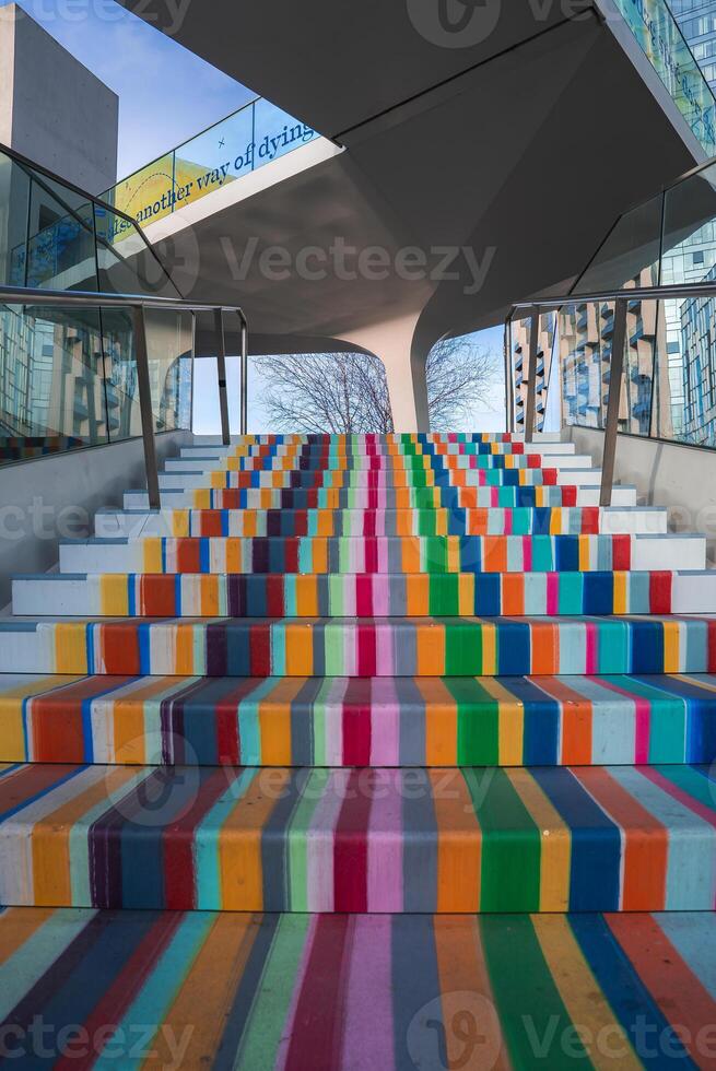 Vibrant staircase with bold striped patterns, glass railings, and a modern structure. A tree and building reflections add depth to the urban scene. photo