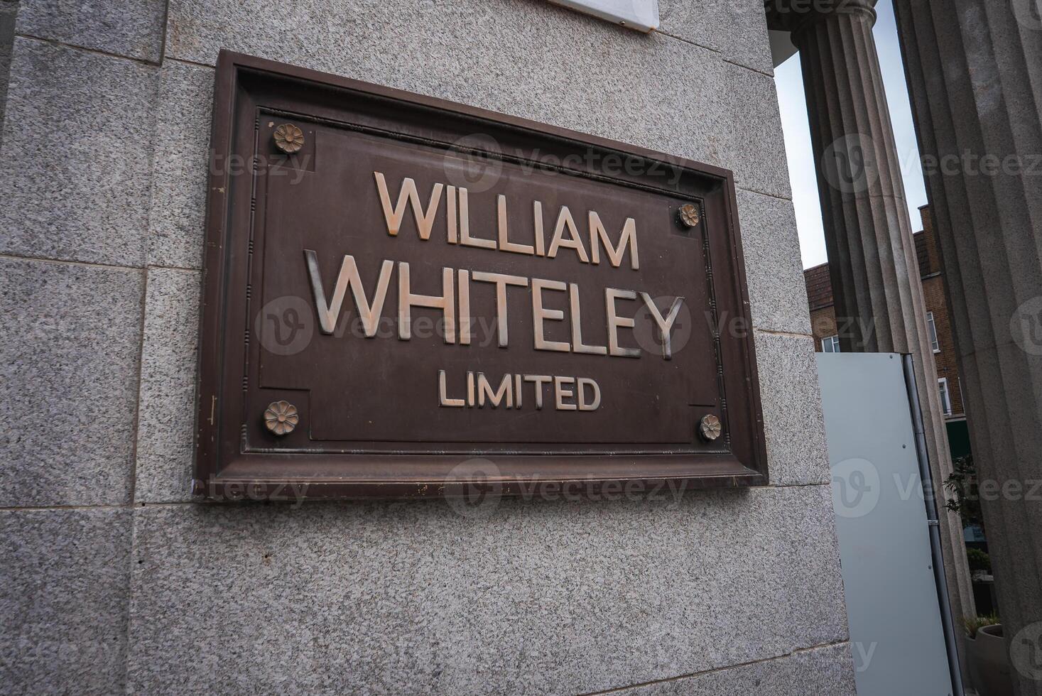 A bronze plaque engraved with William Whiteley Limited is mounted on a light gray stone wall, with part of a column and a building in the background. photo