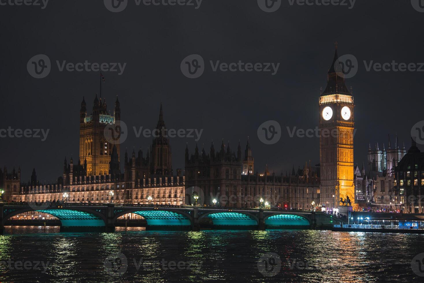 Big Ben and the Houses of Parliament glow against the night sky. Westminster Bridge spans the River Thames, with turquoise lights reflecting on the water. photo