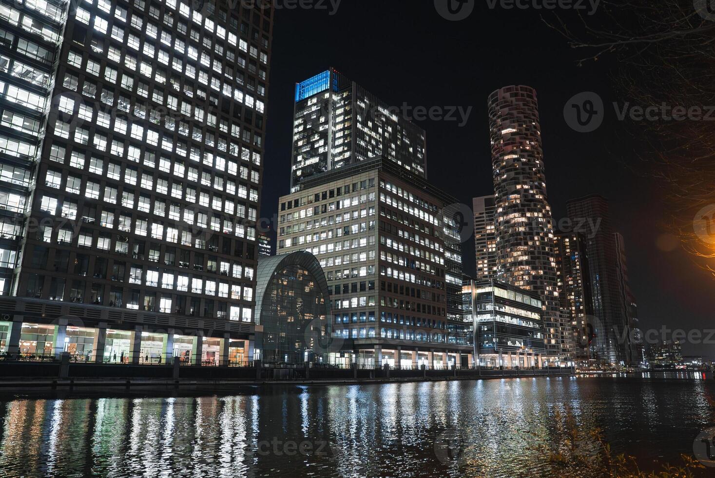 Canary Wharf in London at night, featuring illuminated skyscrapers, the arched Canary Wharf DLR station, and One Park Drive by calm dock waters. photo