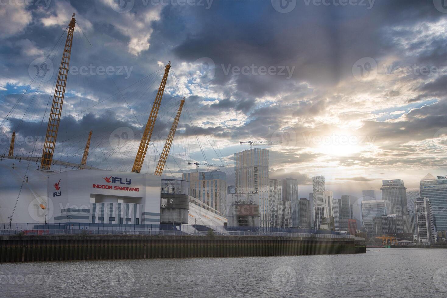 The O2 Arena's white dome and yellow masts stand near the River Thames, with Canary Wharf skyscrapers in the background under a dramatic sky. photo