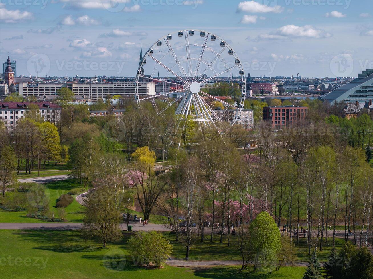 Aerial view of Riga Victory Park in Latvia, featuring a Ferris wheel, blooming sakura trees, green spaces, and the cityscape with the Latvian National Library. photo