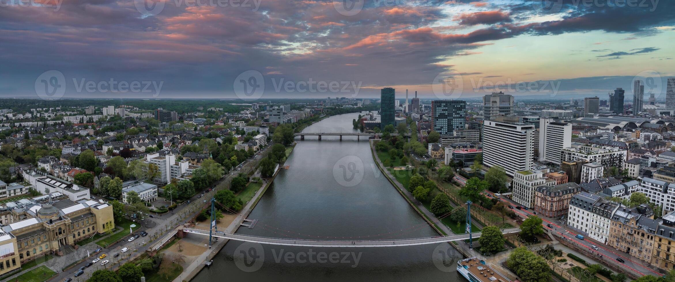 Aerial view of Frankfurt, Germany, featuring the Main River, Eiserner Steg bridge, modern skyline, historic architecture, and a vivid sunset sky. photo
