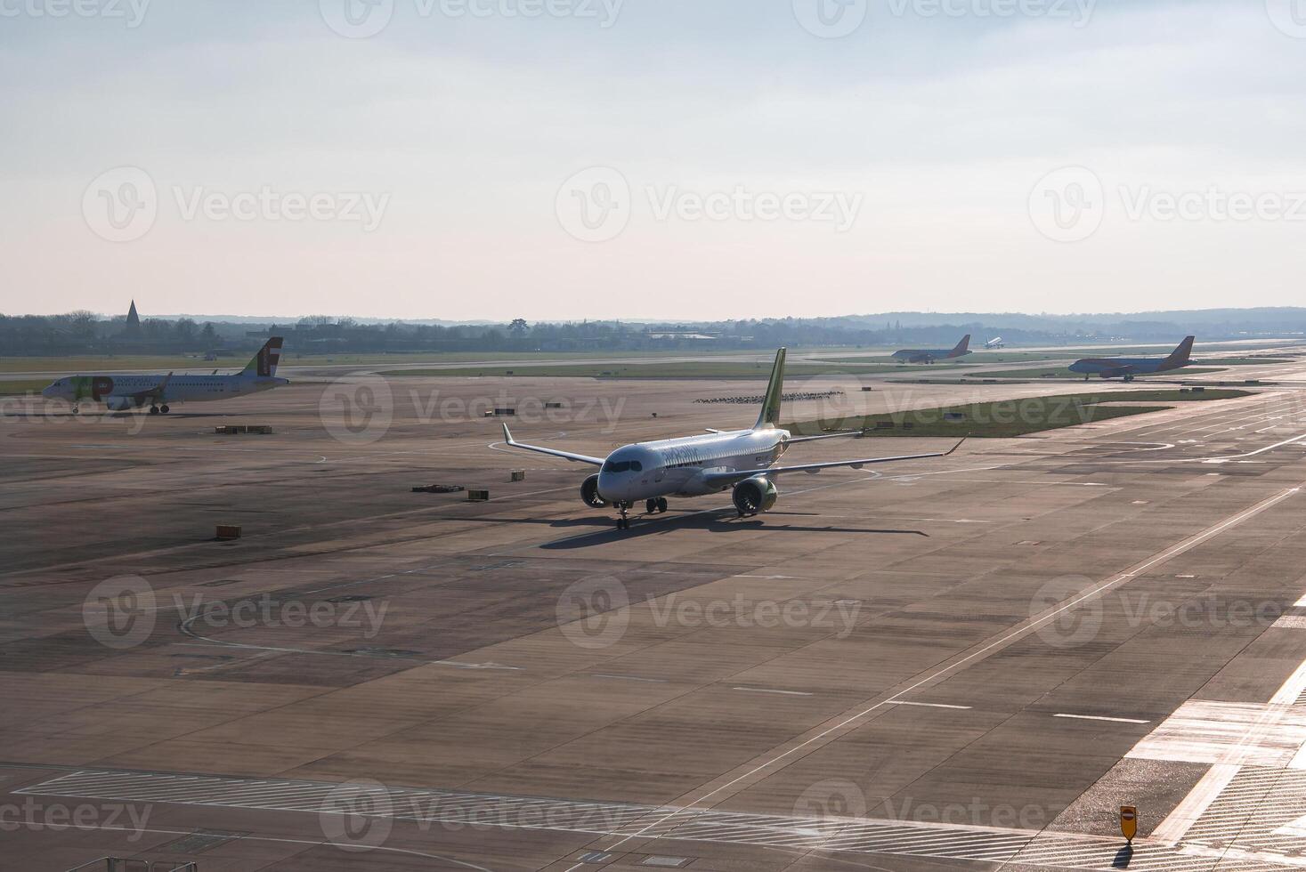 An airport runway with multiple airplanes, including one taxiing in the foreground. A spire or tower is faintly visible on the distant horizon. photo