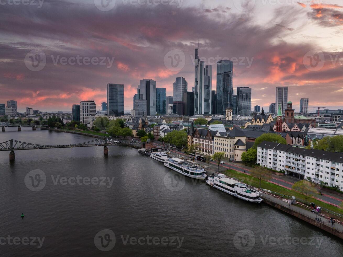 Aerial view of Frankfurt, Germany, featuring the Commerzbank Tower, modern skyscrapers, the Main River, a pedestrian bridge, and a vibrant sunset. photo