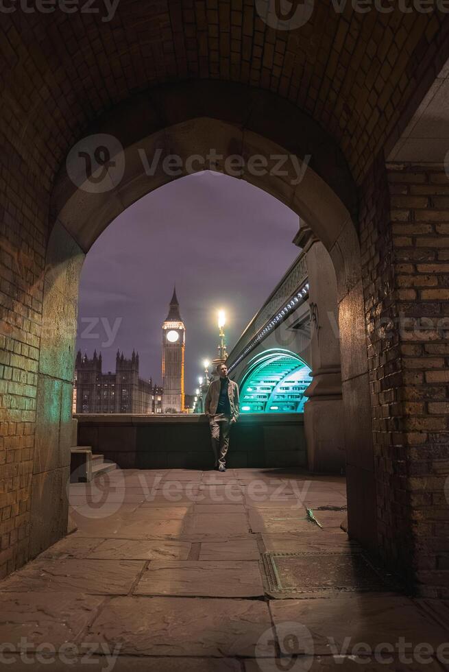 Big Ben and the Houses of Parliament illuminated at night, framed by a brick archway. Westminster Bridge glows to the right, with a person by the railing. photo